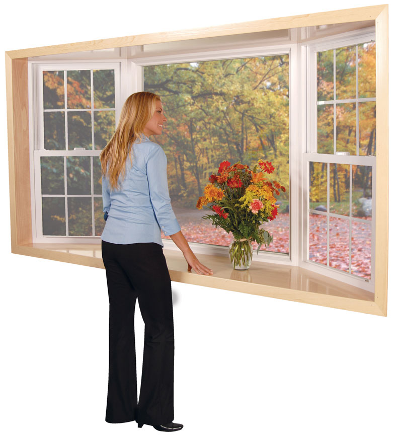 A woman looking away from the camera through a bay window that shows trees and colored leaves on the ground.